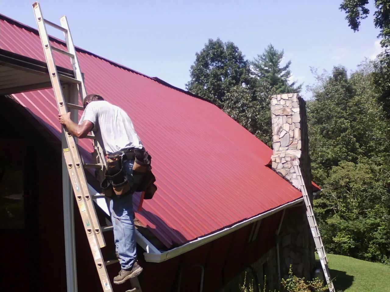 Roofer on a ladder beside a red metal roof and stone chimney in Beryl